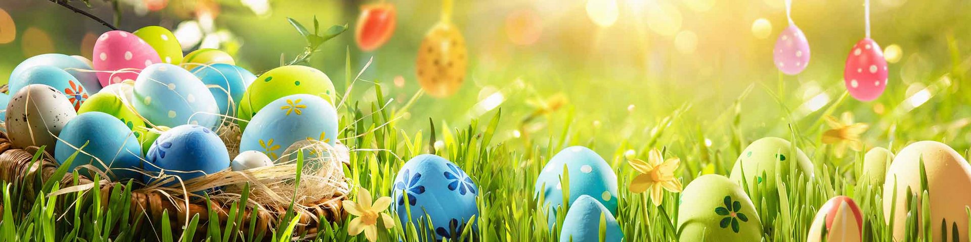 Et kurv fyldt med farverige påskeæg står på frodig grøn græs, omgivet af blomster og sollys. Et par dekorerede æg hænger fra grene i baggrunden. Bildet formidler en festlig og glædelig påskestemning.